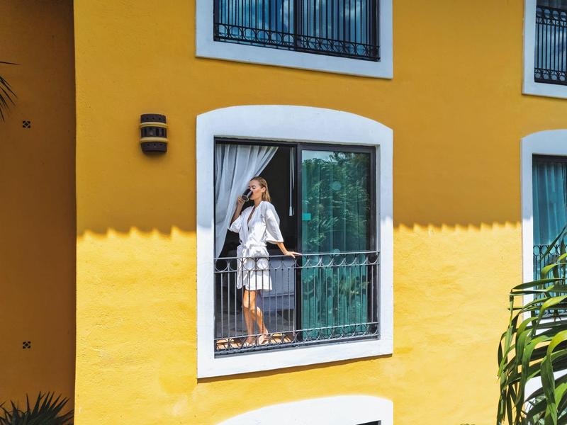 Donna su un balcone di un hotel giallo, circondata da piante verdi.