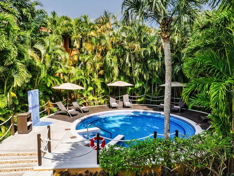 Piscina rotonda con lettini e ombrelloni, circondata da folta vegetazione tropicale.