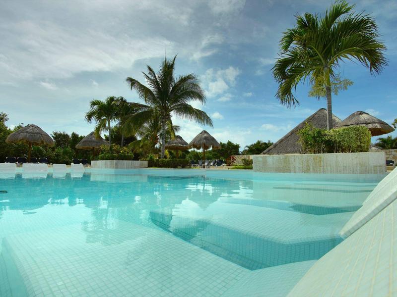 Piscine bleue claire avec palmiers et ciel partiellement nuageux en arrière-plan.