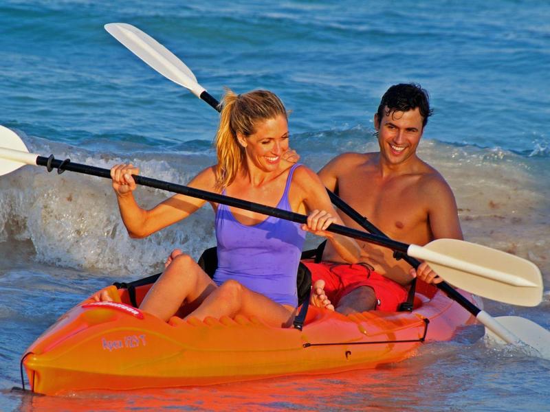 Un couple souriant pagaie en kayak orange près de la plage sur une mer bleue.