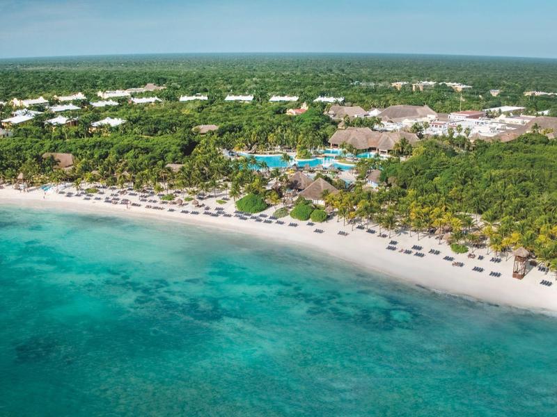 Plage avec sable blanc et eaux turquoise, derrière une forêt dense, grand complexe hôtelier visible.