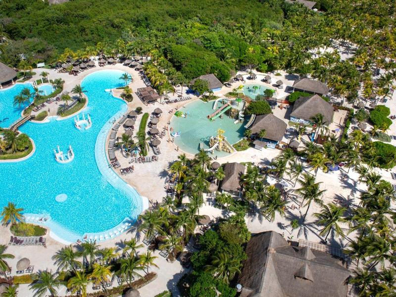 Vue aérienne d'une station balnéaire avec piscine, palmiers et plage de sable blanc.