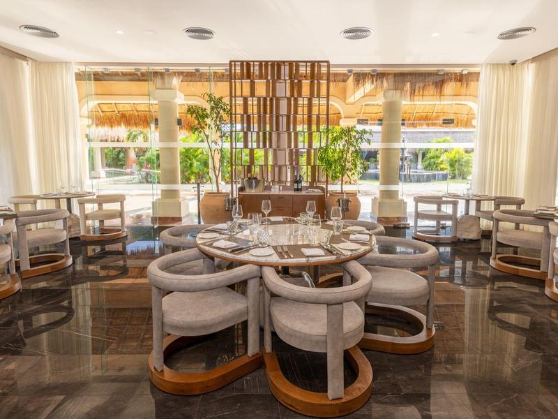 Salle de restaurant élégante avec tables rondes et fauteuils, lumineux grâce aux grandes fenêtres.