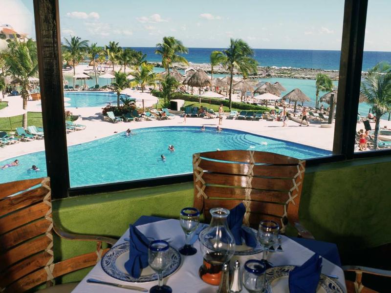 Uitzicht vanaf een gedekte eettafel op een zwembad, palmbomen en de zee op de achtergrond.