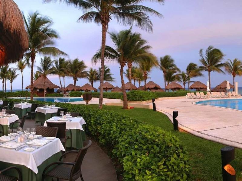 Hotelterras met gedekte tafels naast een groot zwembad en palmbomen bij zonsondergang.