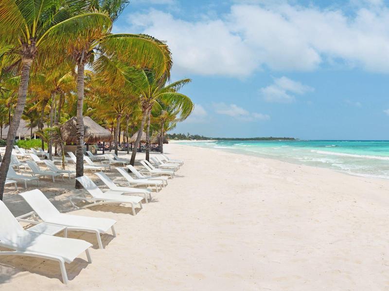 Lettini vuoti sotto palme su una spiaggia di sabbia bianca con oceano turchese e cielo azzurro.