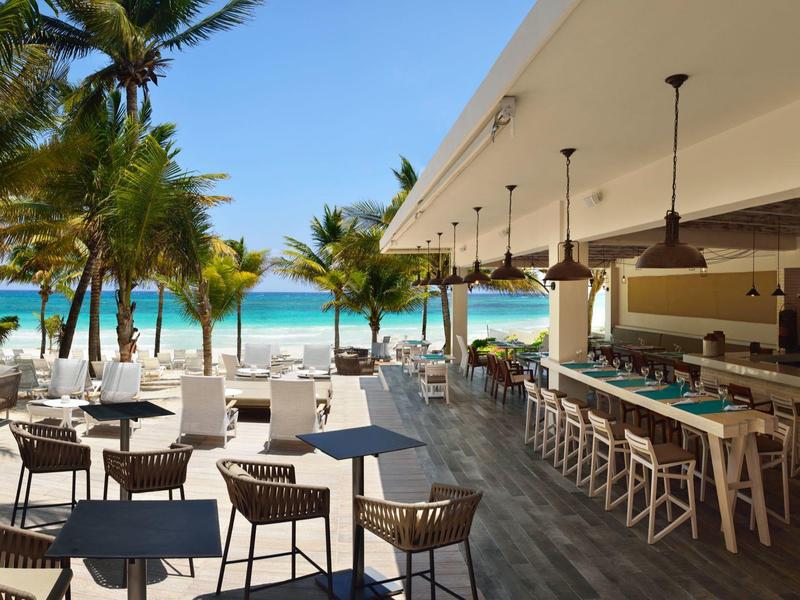 Bar all'aperto sulla spiaggia con posti a sedere sotto una tettoia, palme e vista sull'oceano.