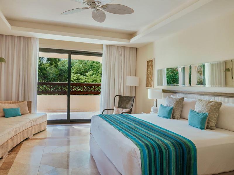 Camera d'hotel luminosa con letto grande, accenti turchesi, divano e balcone con vista sul verde.