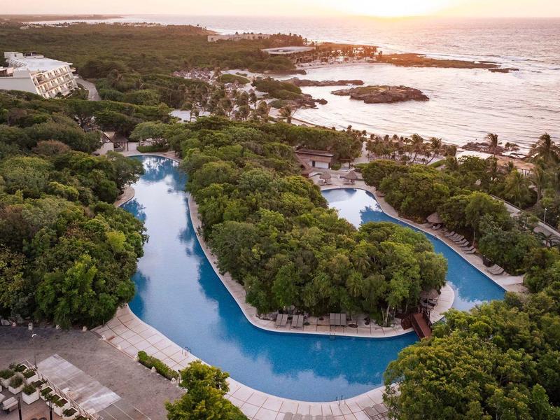 Veduta aerea di un resort tropicale con piscine a laguna circondate da una fitta vegetazione e il mare sullo sfondo.