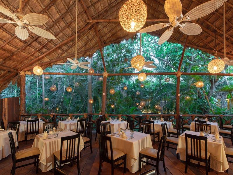 Open-air restaurant with wooden tables, beige cloths, ceiling fans, and lush green surroundings.