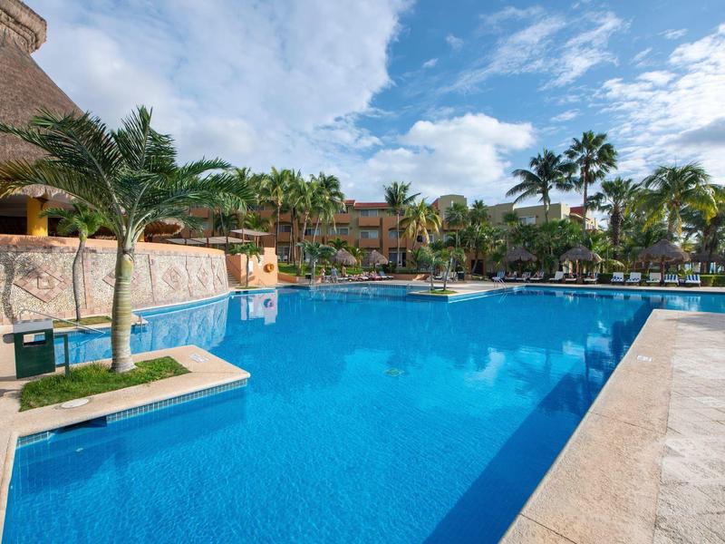 Resort pool surrounded by palm trees and lounge chairs under a partly cloudy sky.