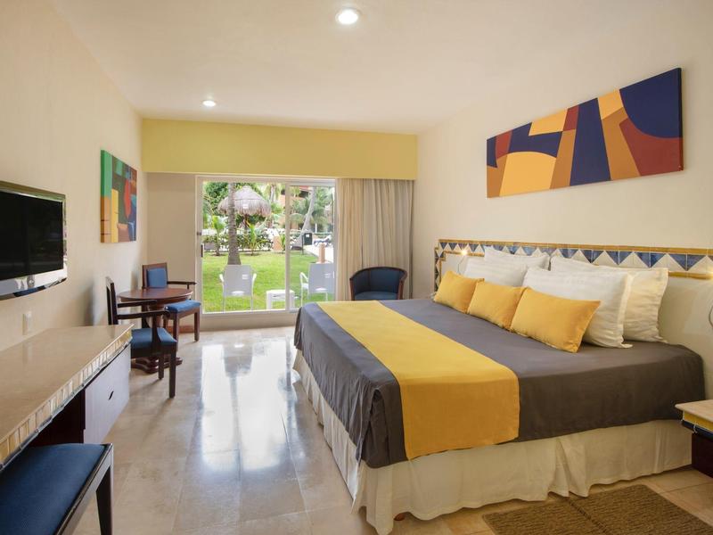 Bright hotel room with large bed, yellow accents, desk, and sliding glass door to garden.