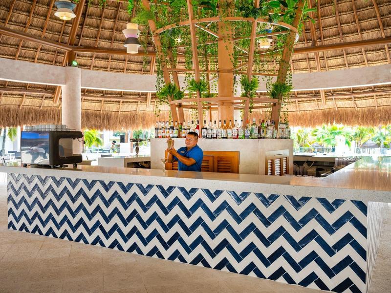 Open-air reception desk with blue zigzag tiles, a thatched roof, and hanging plants in a tropical setting.
