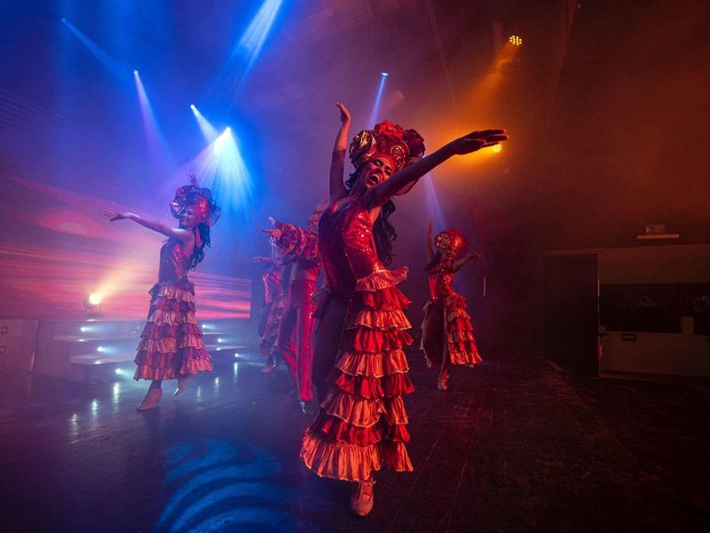 Dancers in colorful flamenco dresses perform on a smoky stage with dramatic blue and orange lighting.
