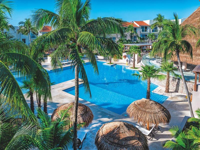 Tropical resort with a large pool, palm trees, and thatched umbrellas under a sunny sky.