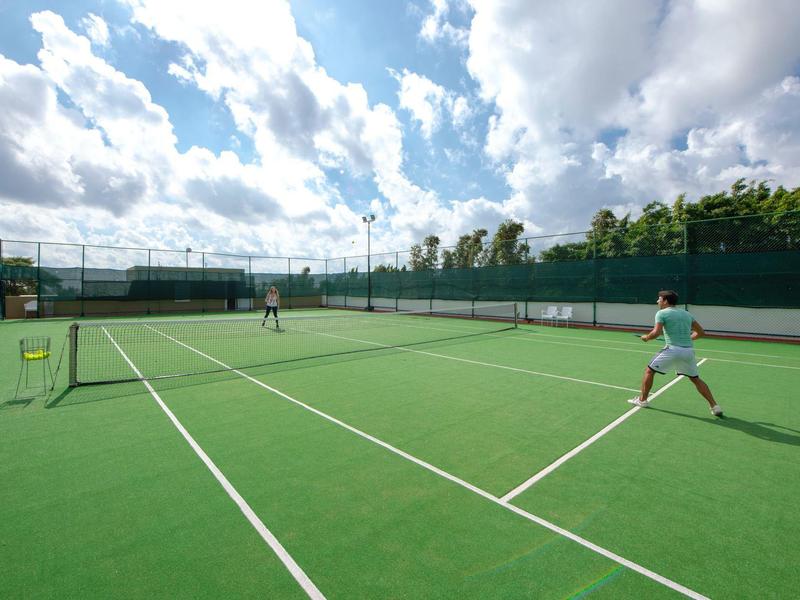 Two people playing tennis on a bright green outdoor court under a partly cloudy sky.