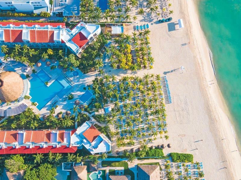 Aerial view of a beachfront resort with a large pool, palm trees, and turquoise ocean water.