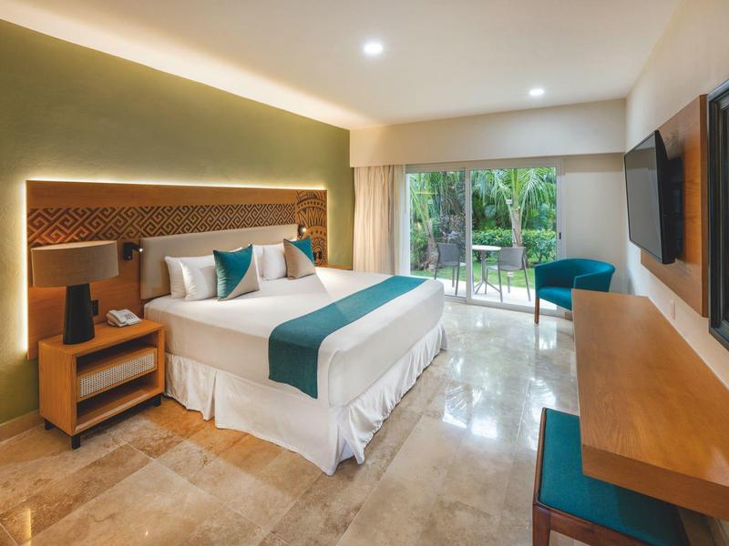 Spacious hotel room with king bed, teal accents, large window overlooking greenery, and modern furnishings.