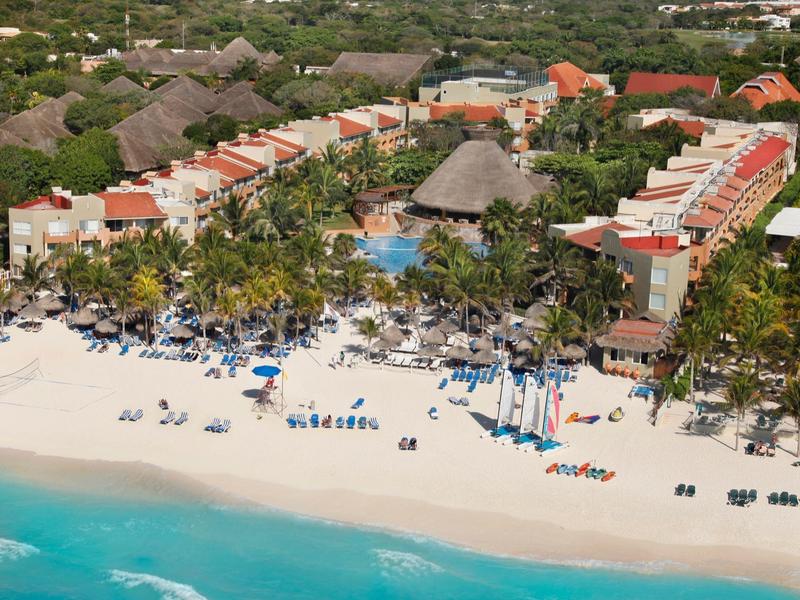 Beachfront resort with turquoise water, white sand, pool, palm trees, and sun loungers under umbrellas.