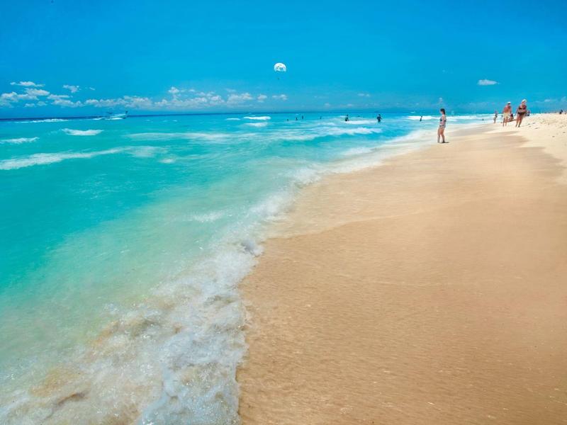 Sandy beach with turquoise waves under a clear blue sky and distant people walking.