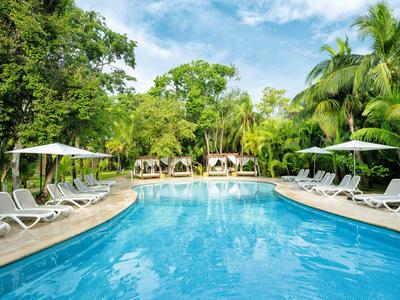 Großer blauer Pool mit Liegestühlen und Sonnenschirmen, umgeben von tropischer Vegetation.