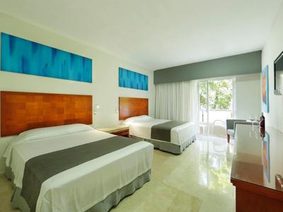 Helles Hotelzimmer mit zwei Doppelbetten, Holzmöbeln, Flachbildschirm und Balkon mit Blick ins Grüne.