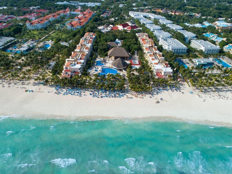 Luftaufnahme eines Strandes mit türkisblauem Wasser, Sandstrand und danebenliegenden Resorts und Palmen.