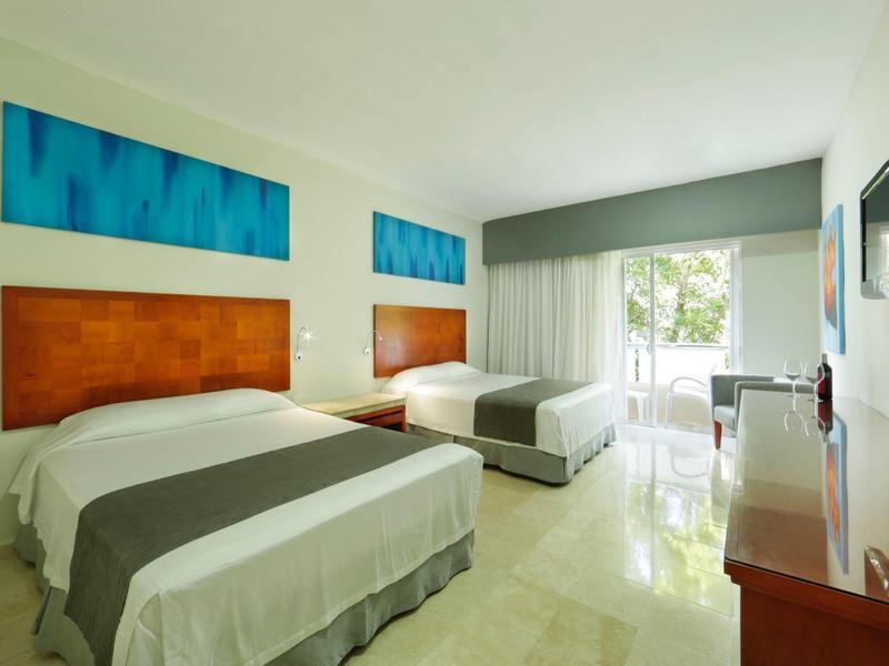 Helles Hotelzimmer mit zwei Doppelbetten, Holzmöbeln, Flachbildschirm und Balkon mit Blick ins Grüne.