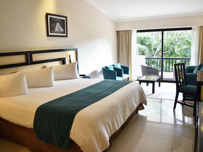 Bright hotel room with large bed, seating area, and view of balcony and greenery.