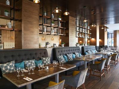 Modern restaurant with wooden tables, comfortable chairs, and elegantly set tables in warm lighting.