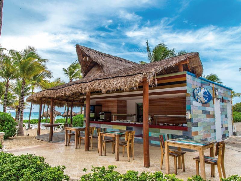 Bar sulla spiaggia con tetto di paglia, tavoli e sedie in legno, palme e cielo blu
