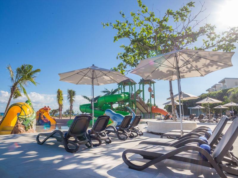 Terrazza solare con lettini, grandi ombrelloni e area giochi colorata per bambini sotto un cielo sereno.
