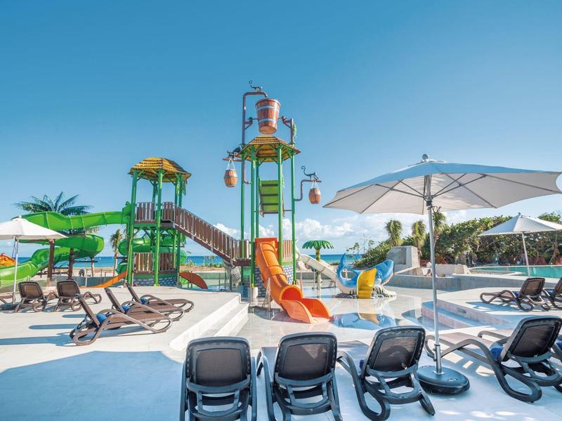 Parco giochi e lettini vicino alla piscina di un hotel sotto un cielo blu limpido.