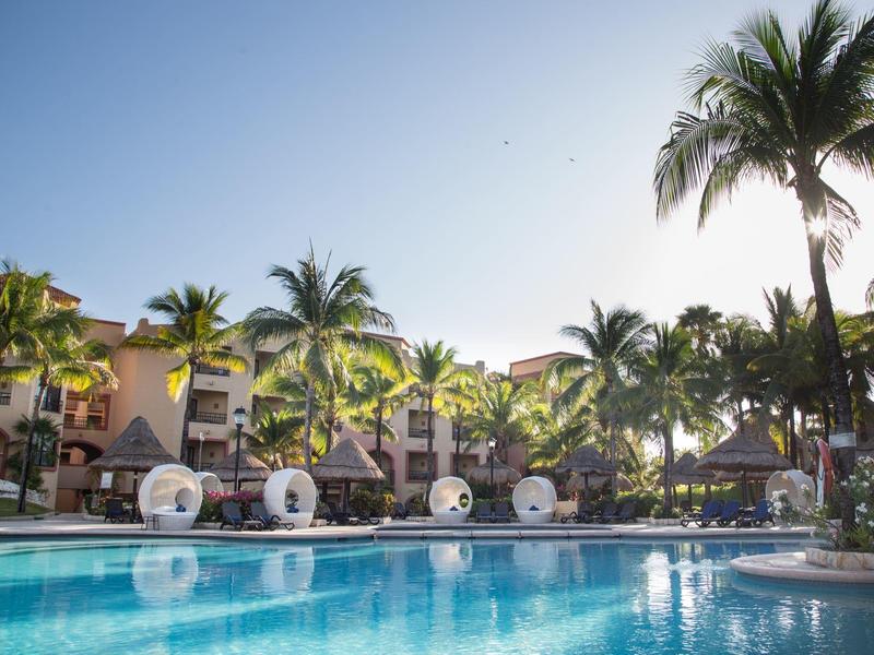 Area piscina con palme e lettini davanti a edifici dell'hotel sotto un cielo sereno.