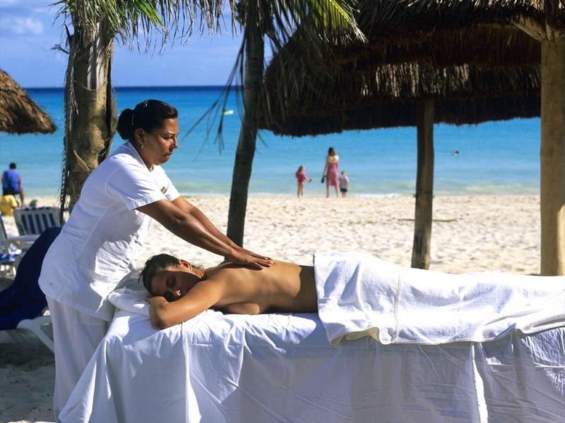 Una donna riceve un massaggio su un lettino da spiaggia sotto palme e tetti di paglia.