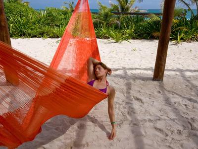 Donna che si rilassa su un'amaca arancione su una spiaggia sabbiosa con vegetazione e oceano sullo sfondo.