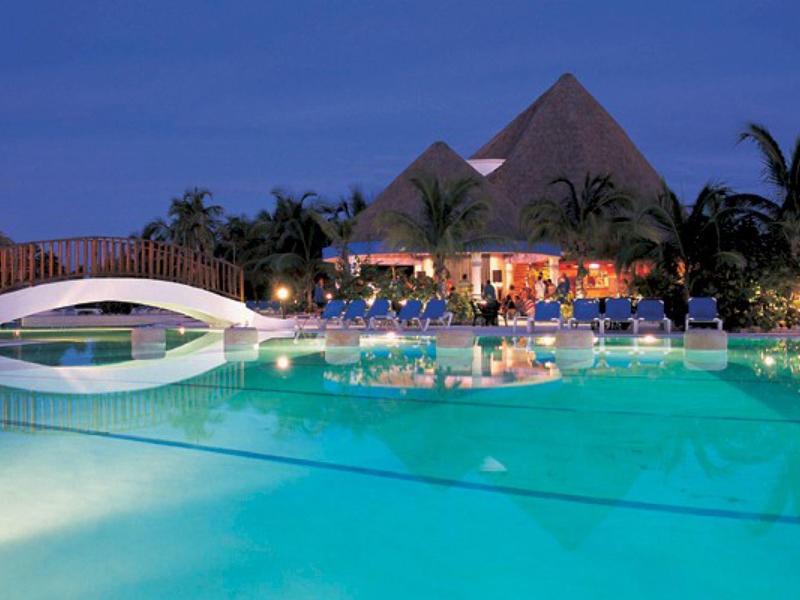 Area piscina del resort con ponte e capanne illuminate di sera.