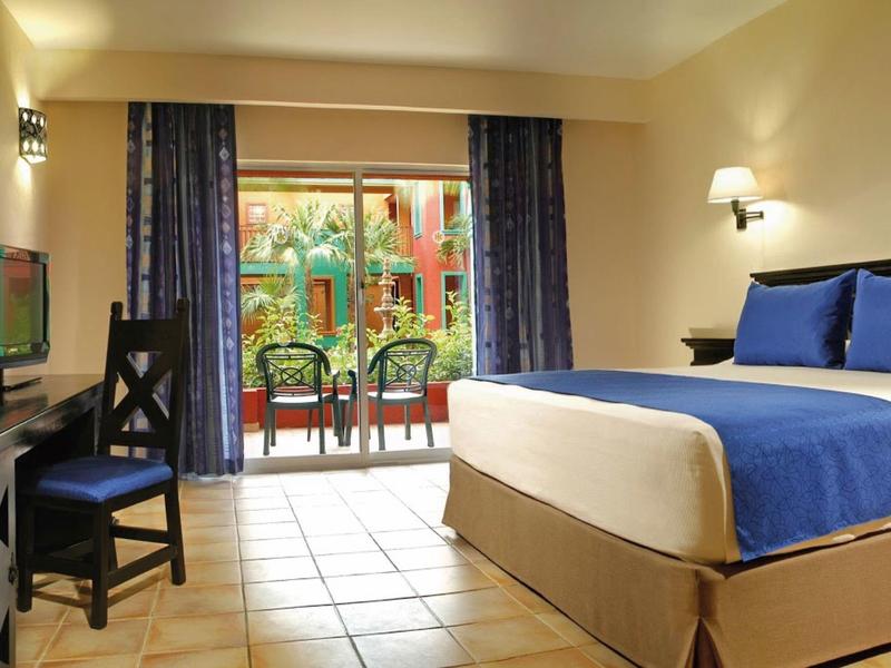 Camera d'hotel con letto queen, arredamento blu, scrivania, TV e balcone con vista giardino.