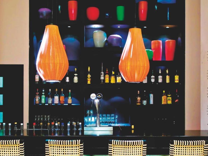 Bar moderno con bottiglie colorate, bicchieri e lampade pendenti arancioni sopra sedie alte.