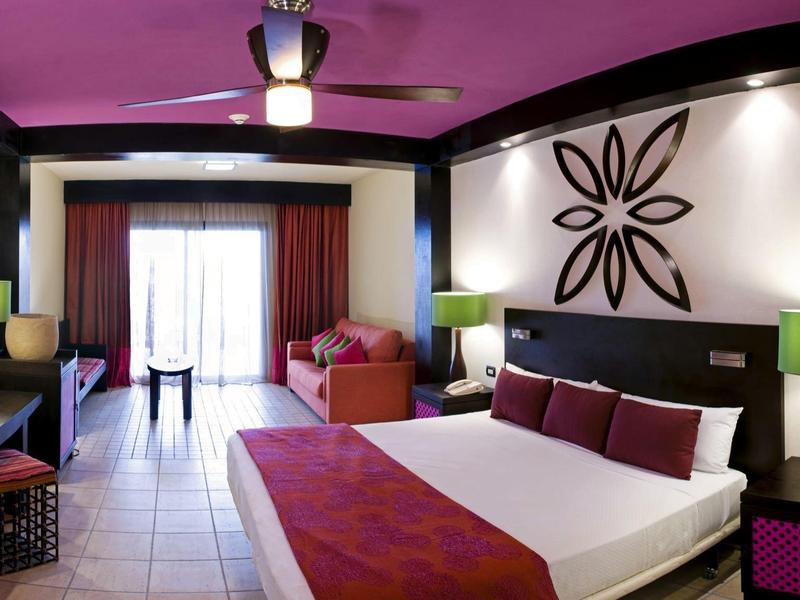 Camera d'hotel moderna con letto matrimoniale, divano, ventilatore a soffitto e accenti viola.