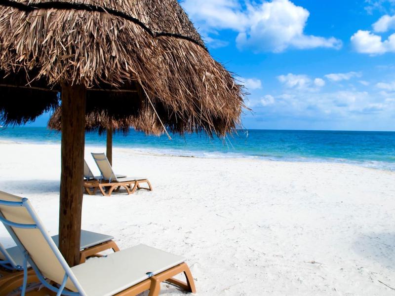Lettini da spiaggia sotto ombrelloni di paglia su una spiaggia di sabbia bianca con cielo azzurro.