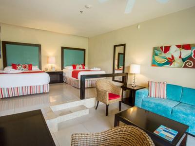 Geräumiges Hotelzimmer mit zwei Doppelbetten, türkisfarbener Couch, Tisch und warmem Licht.