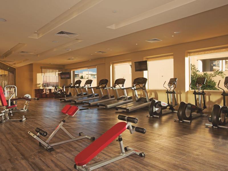 Palestra spaziosa con panche rosse, tapis roulant e ellittiche vicino a grandi finestre.