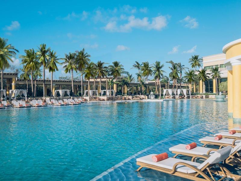 Grande piscina con lettini e palme sotto un cielo azzurro in un resort.