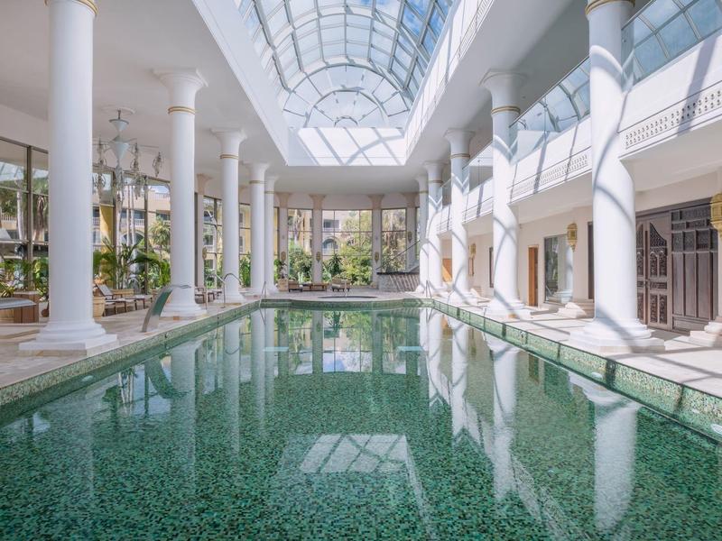 Piscina interna luminosa con colonne, tetto in vetro e terrazza circostante in un hotel.
