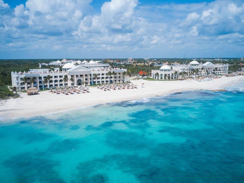 Complesso alberghiero su spiaggia di sabbia bianca con mare turchese sotto un cielo blu.