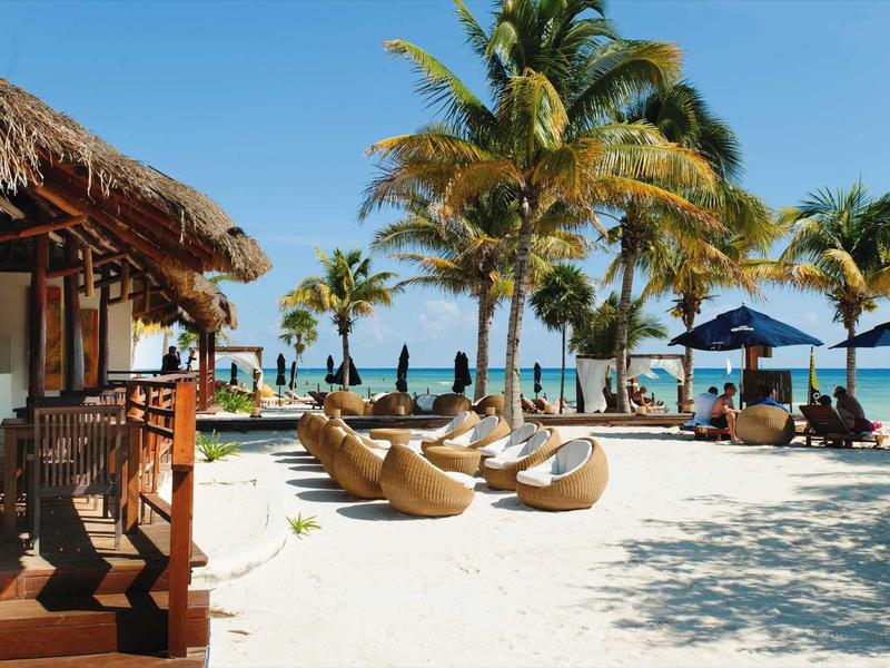 Resort sulla spiaggia con lettini nella sabbia, palme e vista sul mare sotto un cielo limpido.