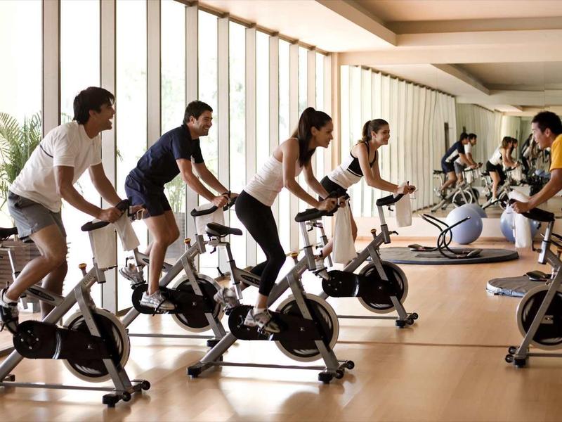 Persone che si allenano insieme su cyclette in una palestra luminosa con grandi finestre.