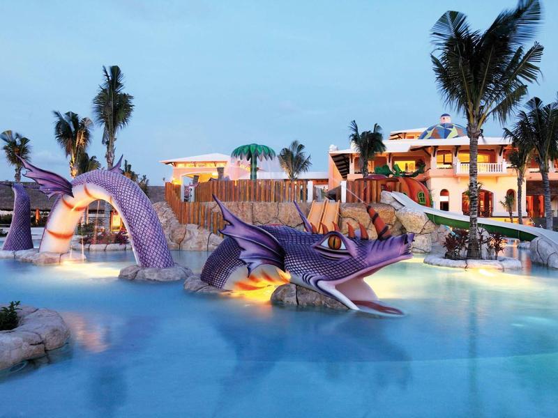 Piscina per bambini con scivoli a forma di drago, circondata da palme e edifici sotto un cielo sereno serale.