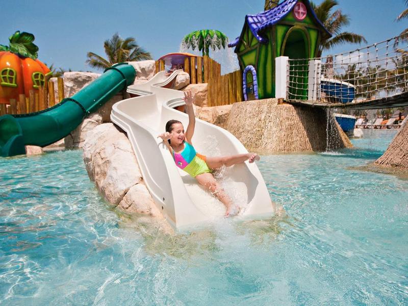 Bambino scivola su uno scivolo d'acqua in un parco acquatico tropicale con case colorate